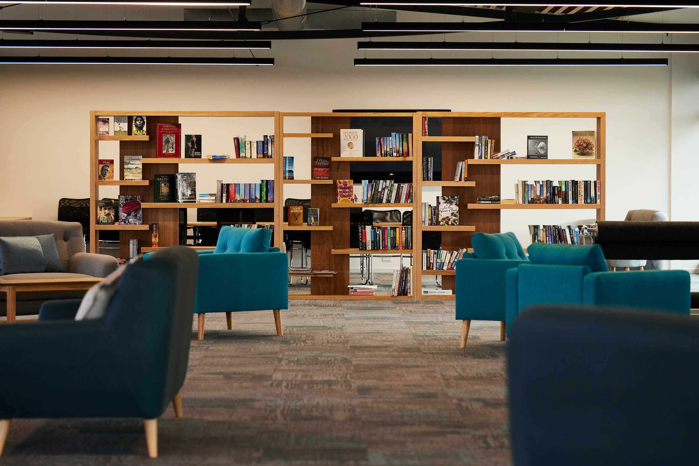 A large open space with teal and grey lounge chairs and a large wooden bookshelf at the rear of the room. The bookshelf is filled with books and the chairs are clustered around tables. Credit: Samuel Shelley.