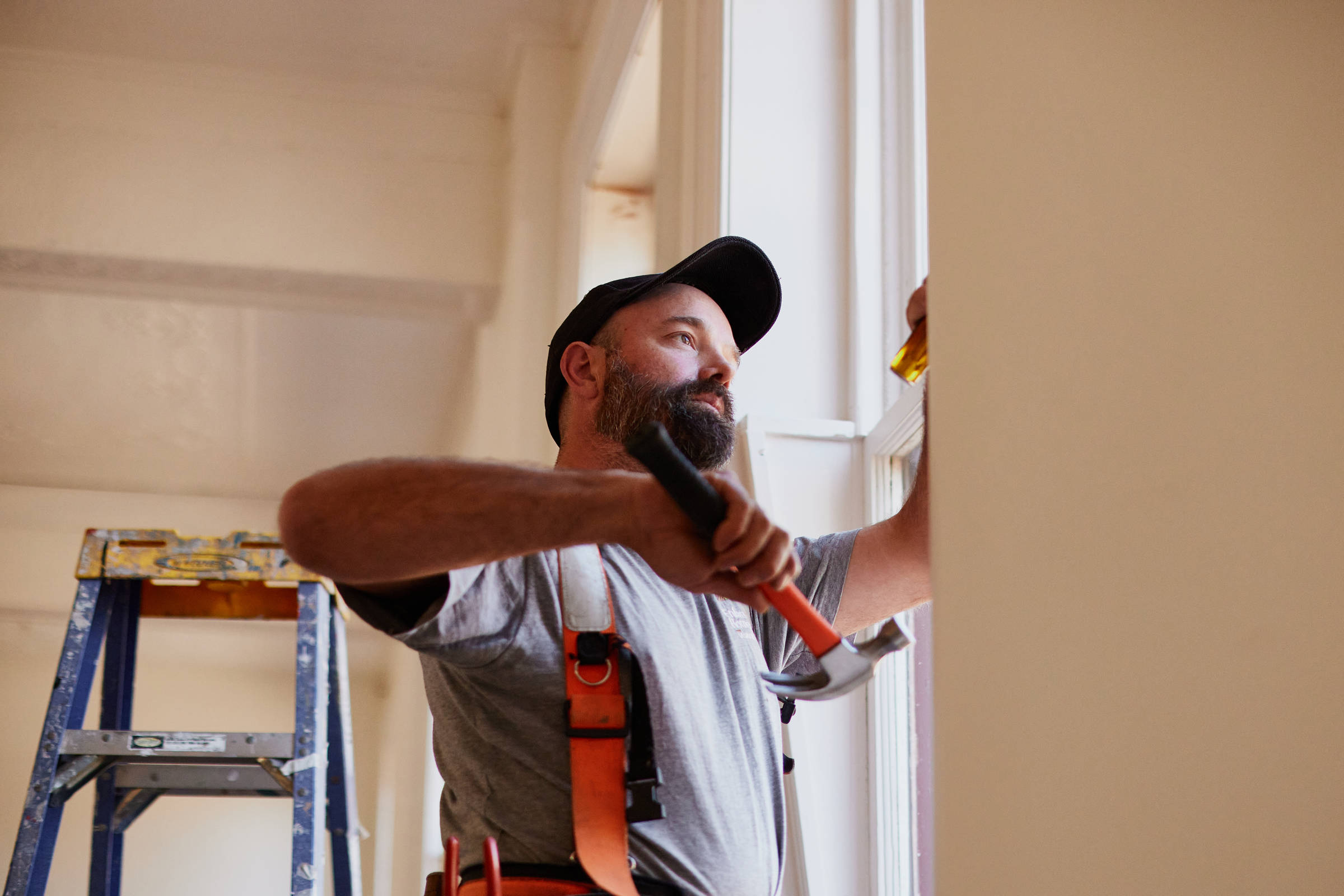 Male carpenter in his 40s who is wearing a toolbelt, looking at a sash window detail with a hammer in his hand and a blue ladder in the background. Credit: Samuel Shelley.