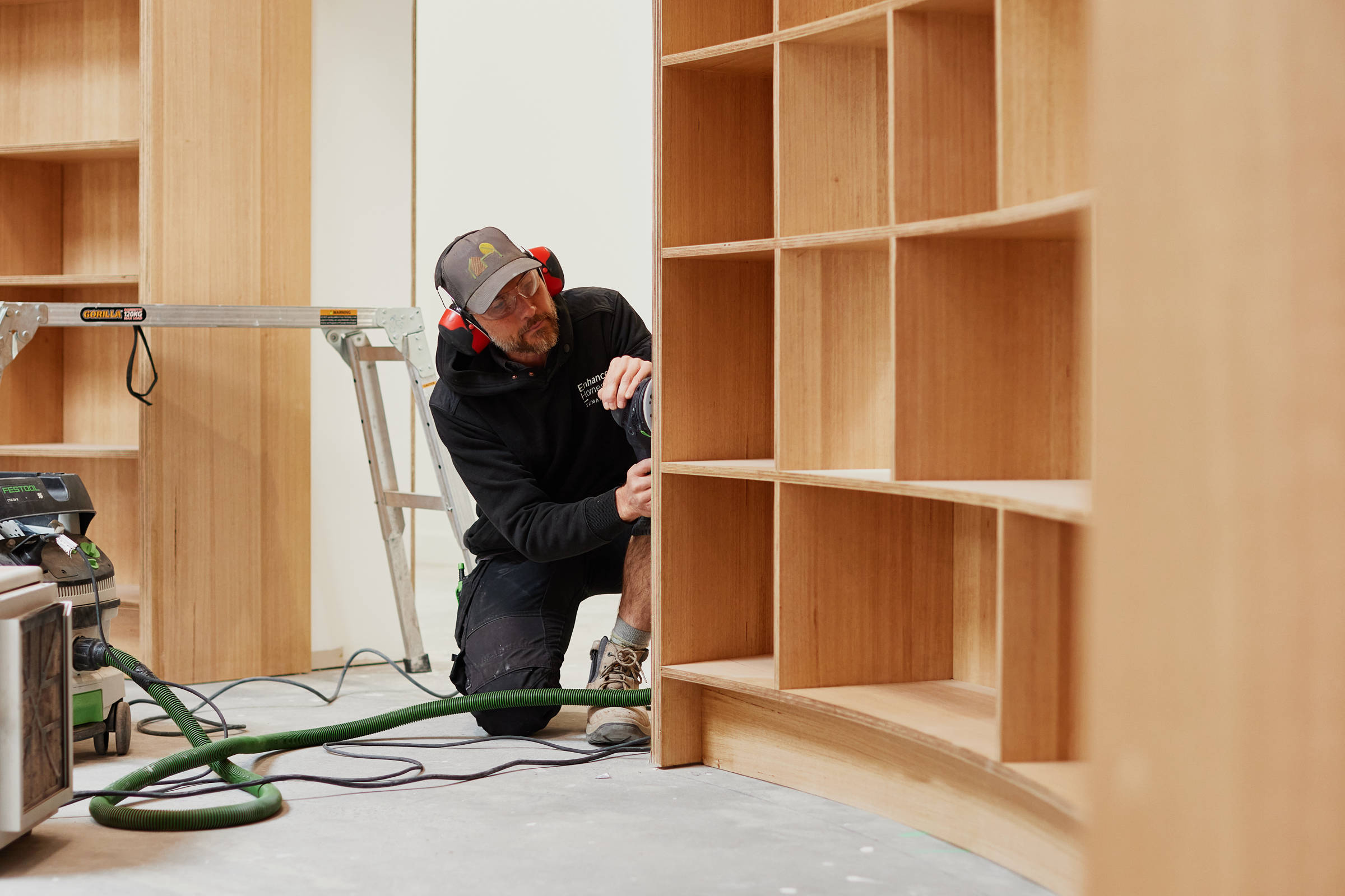 A male carpenter in his late 30s, doing detailed sanding work on the side of curved plywood bookshelves. Credit: Samuel Shelley.