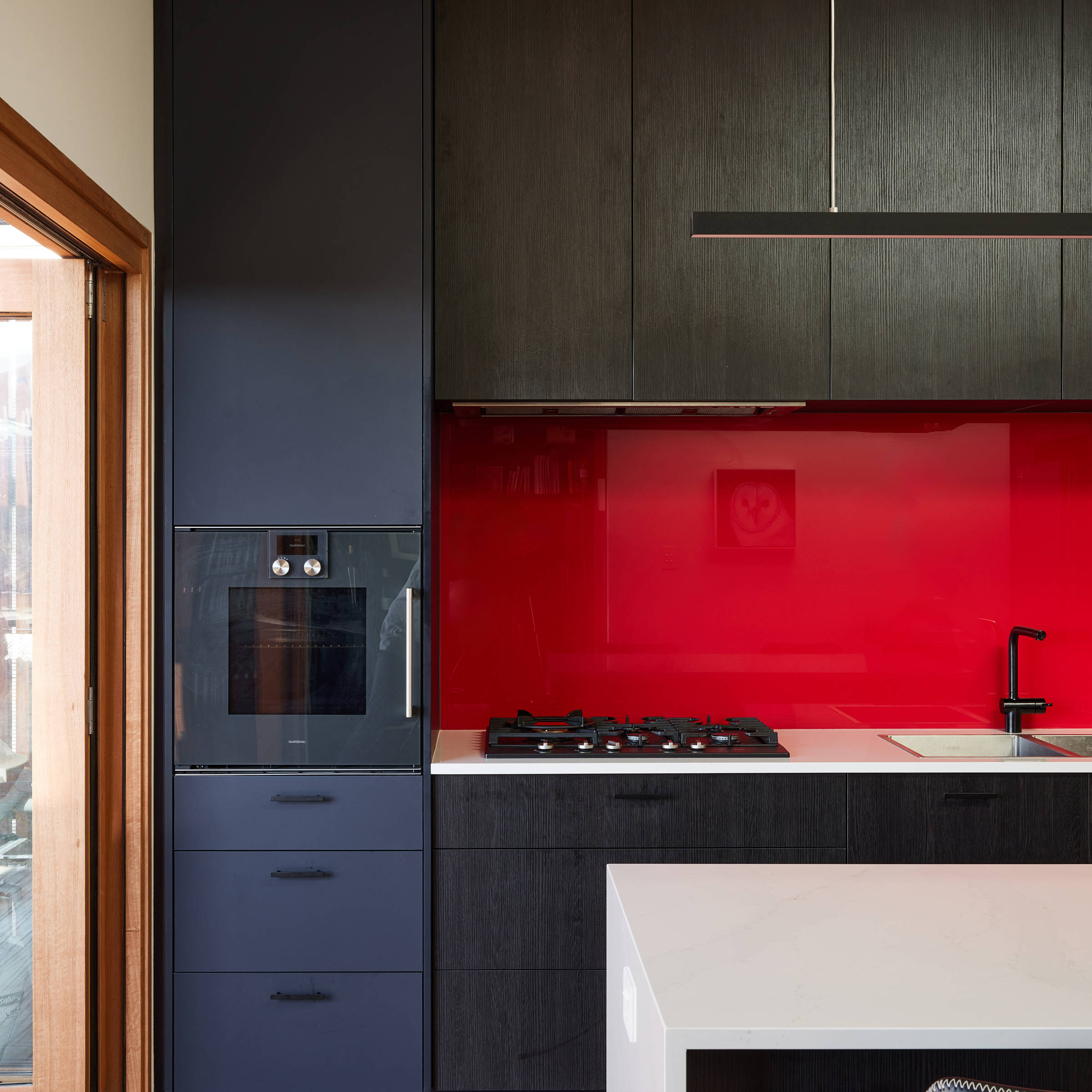 Kitchen leading onto a timber deck through timber framed bifold doors. The kitchen features black cupboards with a visible timber grain, soft grey benchtops and a bright red splashback. The single column floor to ceiling cabinetry to the right of the red splashback is in a dark navy blue. There is an island bench with the same soft grey benchtop. The deck features upright spotted gum narrow timber panels in a symmetrical pattern and a timber table in the same timber tones. Credit: Samuel Shelley.